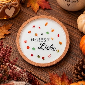 Könnte beinhalten: Ein runder Untersetzer mit Korkrand und weißer Mitte, der den schwarzen Schriftzug "HERBST Liebe" trägt, umgeben von kleinen, bunten Herbstblatt-Illustrationen in Rot-, Orange-, Gelb- und Grüntönen, die kreisförmig angeordnet sind.