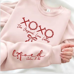 May include: A light pink sweatshirt with red text and graphics. The front features the words "xoxo the Original Love Letters" with nail and wreath graphics. The sleeve displays the word "loved" and the Bible verse John 3:16.