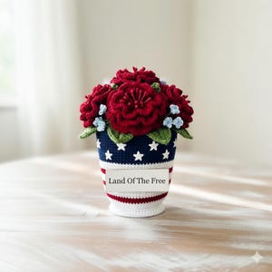 May include: A crocheted floral arrangement in a patriotic theme. The vase is blue with white stars and red and white stripes. The flowers are red with small blue accents and green leaves. The text on the vase reads "Land Of The Free."