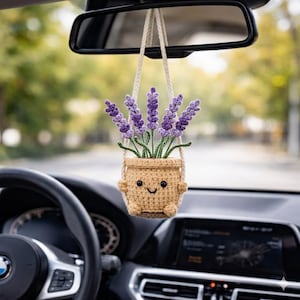 May include: A crocheted car air freshener featuring a smiling pot with lavender flowers. The pot is beige with a smiling face and small arms. The lavender flowers are purple and green. The air freshener hangs from a string attached to the car's rearview mirror.