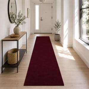 Puede incluir: Una alfombra de pasillo larga y rectangular de color burdeos profundo y sólido. La alfombra presenta una textura suave y uniforme, colocada centralmente sobre un suelo de madera clara, extendiéndose desde el frente hacia una puerta blanca.
