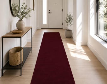 Puede incluir: Una alfombra de pasillo larga y rectangular de color burdeos profundo y sólido. La alfombra presenta una textura suave y uniforme, colocada centralmente sobre un suelo de madera clara, extendiéndose desde el frente hacia una puerta blanca.