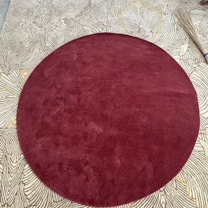May include: A solid deep burgundy circular rug with a plush texture. The rug is placed on top of a larger rectangular rug featuring a cream and beige background with a repeating line-drawn floral pattern in white and gold thread.