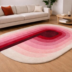Luxury hand-tufted silk oval area rug in a Scandinavian living room with oak hardwood flooring. Features a bold gradient "P" arch motif in ombre shades of burgundy, crimson, rose pink, and blush ivory. Styled with a cream linen sofa, terracotta accent pillow, and natural wood coffee table — a versatile statement piece for contemporary interiors.