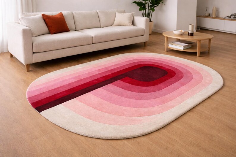 Luxury hand-tufted silk oval area rug in a Scandinavian living room with oak hardwood flooring. Features a bold gradient "P" arch motif in ombre shades of burgundy, crimson, rose pink, and blush ivory. Styled with a cream linen sofa, terracotta accent pillow, and natural wood coffee table — a versatile statement piece for contemporary interiors.