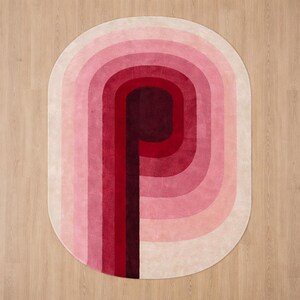 Luxury hand-tufted silk area rug in an oval shape featuring a bold gradient "P" letter motif in ombre shades of deep burgundy, crimson red, and blush pink on an ivory background, photographed from above on light wood flooring. Modern designer rug available in 6x9 and 8x10 sizes.