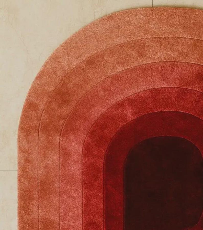 Close-up of a luxury hand-tufted silk area rug featuring concentric arch gradient bands transitioning from soft terracotta and dusty rose to deep crimson and rich burgundy. Plush velvety pile texture and precision tufting craftsmanship are clearly visible, highlighting exceptional silk quality and smooth ombre color gradation.