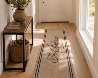 Puede incluir: Una alfombra rectangular de color beige con un borde de doble línea marrón oscuro y el texto The Andersens escrito en cursiva negra con un adorno decorativo. La alfombra está colocada sobre un suelo de madera clara en un pasillo.