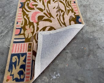 Puede incluir: Una alfombra rectangular con un patrón repetitivo de formas florales y geométricas en verde oliva, rosa suave y azul marino sobre un fondo crema, con una esquina doblada que revela un reverso de tela gris claro y bordes rematados.