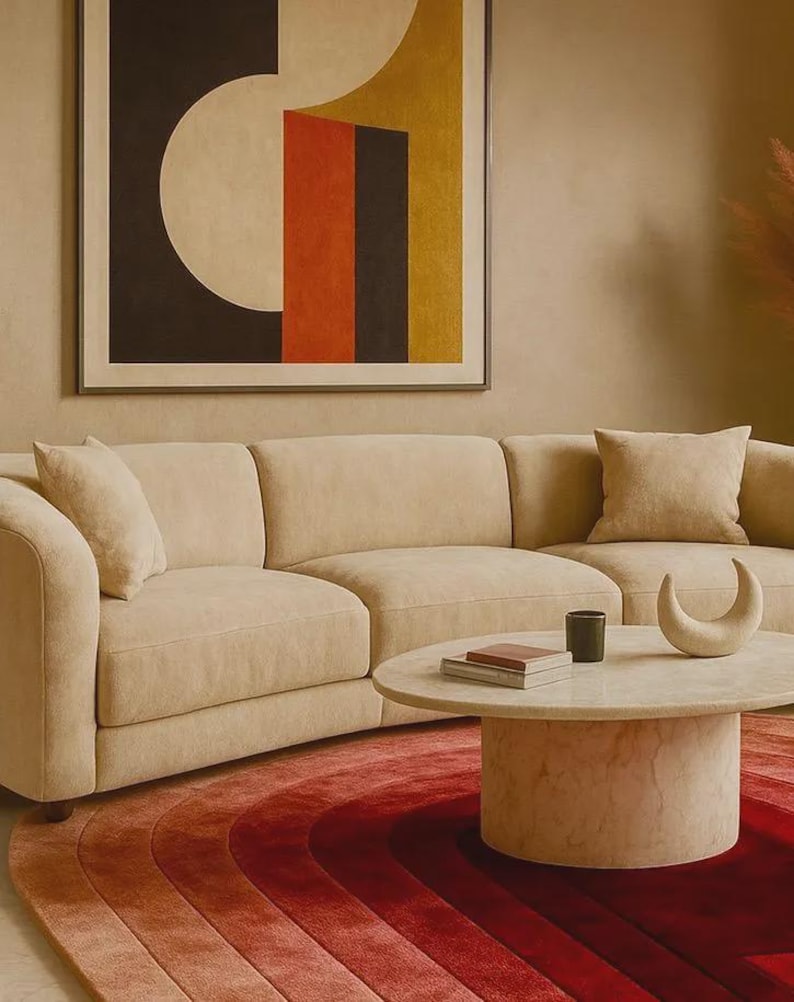 Close-up detail of a luxury hand-tufted silk gradient area rug in rich ombre tones of deep burgundy, crimson red, and terracotta rose, styled beneath a round travertine marble coffee table and a curved cream bouclé sectional sofa. The concentric arch pattern and plush pile texture are prominently visible, complemented by a bold mid-century modern wall art in the background. Ideal statement rug for warm-toned contemporary living spaces. Available in 6x9 and 8x10.