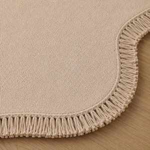 May include: A beige rug with a textured, pebbled surface and a curved edge. The perimeter of the rug is finished with a dense, matching beige fringe trim that follows the rounded contour of the textile piece, creating a uniform appearance.