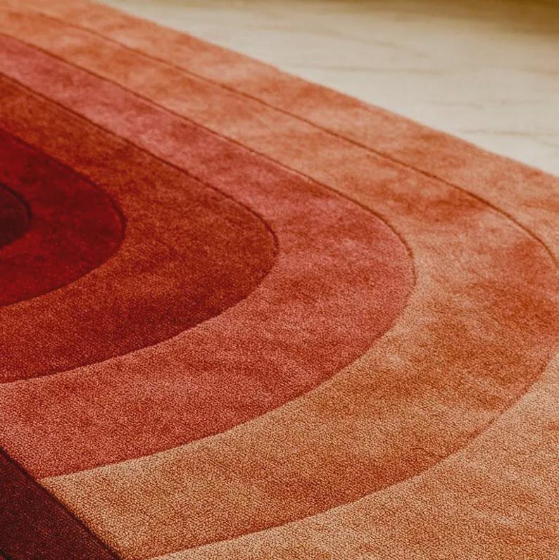 Low-angle close-up of a luxury hand-tufted silk area rug shot from floor level, revealing the curved edge profile and plush pile depth. The gradient ombre pattern flows in sweeping concentric arches from warm terracotta and burnt sienna on the outer edges to deep crimson and dark burgundy at the center. The angled perspective highlights the silk sheen, even pile height, and crisp band definition of the hand-tufted craftsmanship, set against a light marble floor background.
