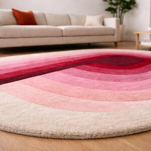 Ground-level close-up of a luxury hand-tufted silk oval area rug highlighting the thick plush pile and neatly finished curved edge. Ombre gradient arches flow from cream ivory through blush and rose pink to deep burgundy at the center. Silk sheen and dense pile depth captured beautifully, with a Scandinavian living room softly blurred in the background.