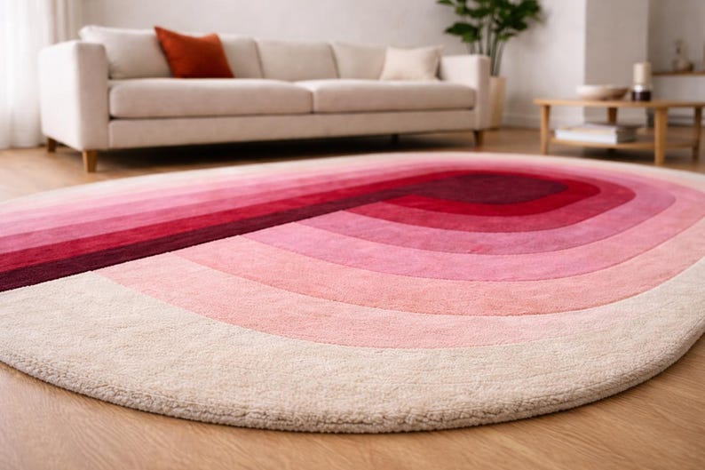 Ground-level close-up of a luxury hand-tufted silk oval area rug highlighting the thick plush pile and neatly finished curved edge. Ombre gradient arches flow from cream ivory through blush and rose pink to deep burgundy at the center. Silk sheen and dense pile depth captured beautifully, with a Scandinavian living room softly blurred in the background.