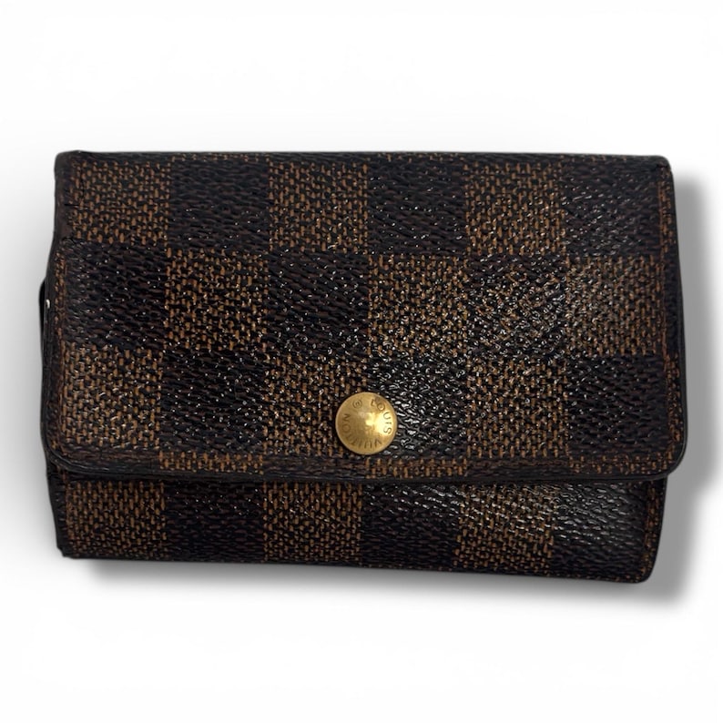 May include: A brown and black checkered wallet with a gold button closure. The wallet is rectangular and has a flap that folds over to close. The pattern is a repeating grid of dark brown and black squares.