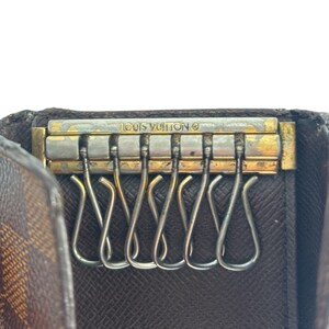 May include: Close-up of a brown and tan Louis Vuitton key holder. The key holder features a gold-toned metal bar with the brand name and six silver key hooks. The interior is a dark brown leather.