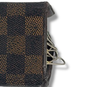 May include: Close-up of a brown and tan checkered key holder. The key holder is made of leather and has a metal key ring attached. The key holder is designed to hold multiple keys. The key holder is a small, compact accessory.