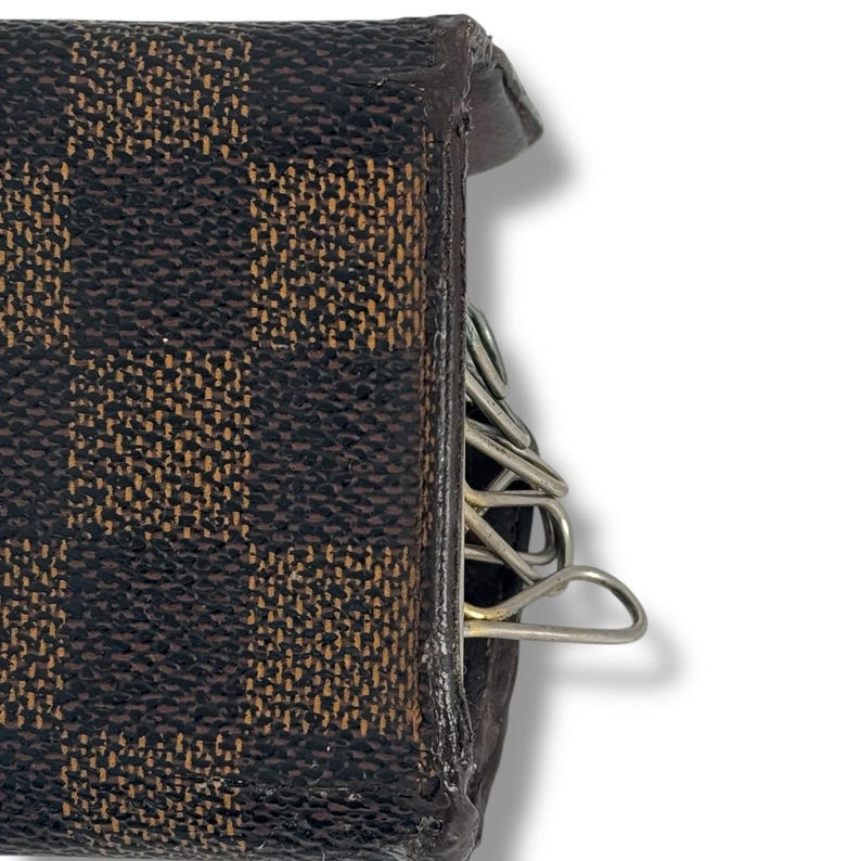 May include: Close-up of a brown and tan checkered key holder. The key holder is made of leather and has a metal key ring attached. The key holder is designed to hold multiple keys. The key holder is a small, compact accessory.