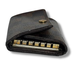 May include: A dark brown leather key holder with a snap closure. The key holder is open, revealing several metal key hooks. The brand name is visible on the metal bar.