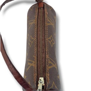 May include: A brown cylindrical handbag with a zip closure and a brown leather strap. The bag features a repeating pattern of interlocking letters in a lighter brown colour. The zip is gold-coloured.