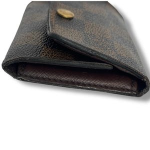 May include: Brown and tan checkered wallet with a gold-colored button closure. The wallet is made of a textured material and has a dark brown interior. The wallet is closed and angled towards the viewer.