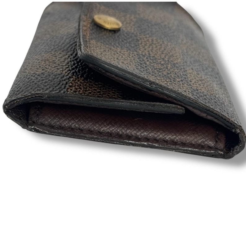 May include: Brown and tan checkered wallet with a gold-colored button closure. The wallet is made of a textured material and has a dark brown interior. The wallet is closed and angled towards the viewer.