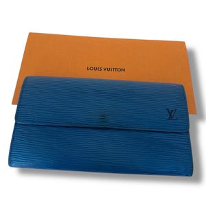 May include: A blue leather wallet with a textured surface and a flap closure. The wallet features the Louis Vuitton logo and is displayed with an orange box that has the words "LOUIS VUITTON" printed on it.