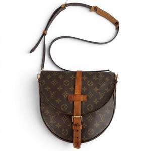 May include: A brown and dark brown monogram patterned shoulder bag with a rounded bottom shape. The bag features a vertical tan leather strap with a metal buckle closure at the center and an adjustable shoulder strap with a tan leather shoulder pad.