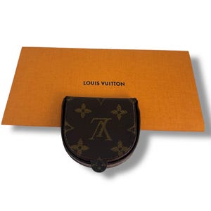 May include: A brown and tan coin purse with the Louis Vuitton logo and a matching orange box with the brand name printed on it. The coin purse has a curved shape and a snap closure.