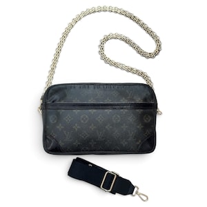 May include: A rectangular black shoulder bag featuring a dark gray monogram pattern, black leather trim, and a long gold-tone chain strap. A separate black fabric adjustable shoulder strap with gold-tone metal hardware is positioned below the bag.