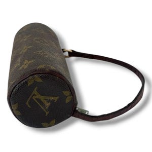 May include: Cylindrical brown handbag with a dark brown handle. The bag has a repeating pattern of brown logos on a lighter brown background. The ends of the bag are a darker brown. A small silver zip is visible.