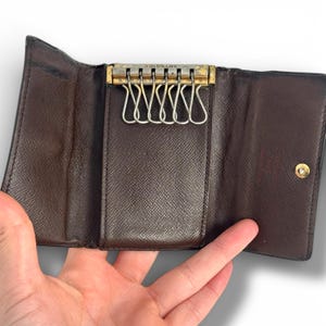 May include: Open brown leather key holder with a gold-toned metal key ring holder. The key holder is open, revealing multiple key hooks. The interior is a lighter shade of brown. The key holder is held open by a hand.