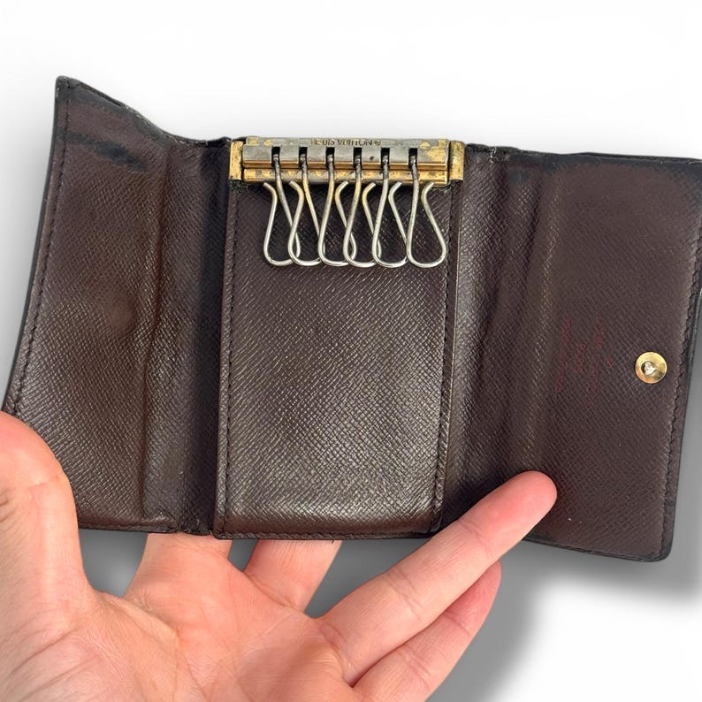 May include: Open brown leather key holder with a gold-toned metal key ring holder. The key holder is open, revealing multiple key hooks. The interior is a lighter shade of brown. The key holder is held open by a hand.