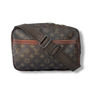 May include: Brown crossbody bag with a repeating logo pattern. The bag features a brown adjustable strap and a brown leather accent. The bag has a rectangular shape and a zip closure.