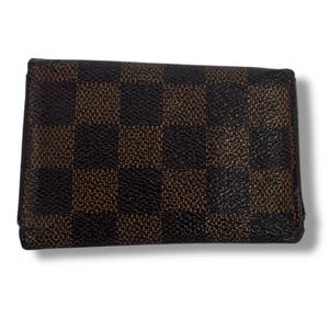 May include: A brown and black checkered wallet. The wallet has a rectangular shape with a repeating pattern of dark brown and black squares. The wallet is closed and appears to be made of leather or a similar material.