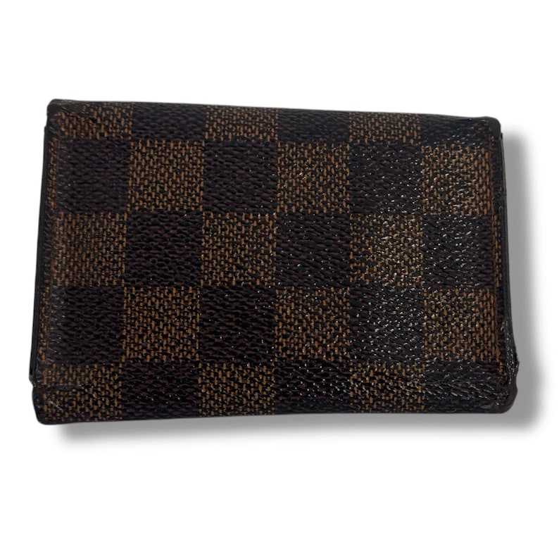 May include: A brown and black checkered wallet. The wallet has a rectangular shape with a repeating pattern of dark brown and black squares. The wallet is closed and appears to be made of leather or a similar material.