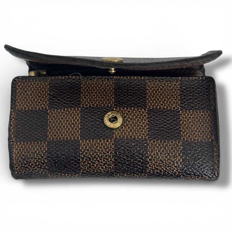 May include: A brown and black checkered pattern key holder with a gold-tone button closure. The key holder is open, revealing the interior. The design is a classic, recognizable pattern.