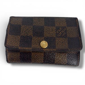 May include: A brown and black checkered wallet with a gold button closure. The wallet has a rectangular shape and a textured surface. The brand name is visible on the front.