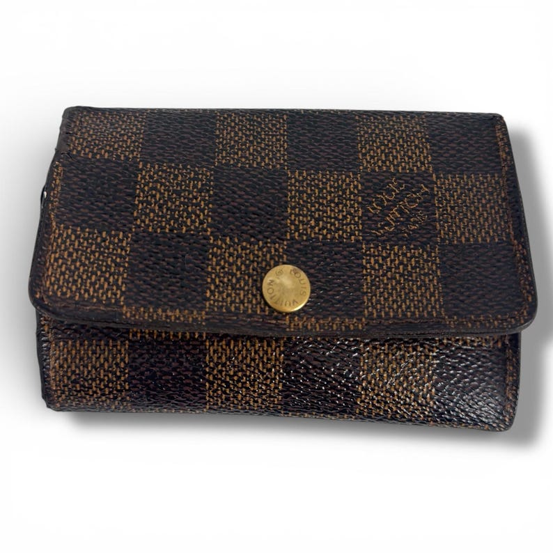 May include: A brown and black checkered wallet with a gold button closure. The wallet has a rectangular shape and a textured surface. The brand name is visible on the front.