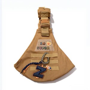Könnte beinhalten: Beigefarbene Haustiertasche mit brauner Schnalle und Riemen. Die Tasche trägt den Text "IM SUPER" und einen dekorativen "Z"-Anhänger mit einer Cartoonfigur. Enthält einen Karabinerhaken und ein blau-oranges Seil.