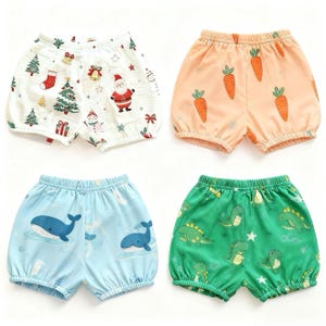 May include: Four pairs of baby shorts with elasticated waistbands. One pair is white with Christmas patterns, another is peach with carrot designs, a third is light blue with whale illustrations, and the last is green with dinosaur graphics.