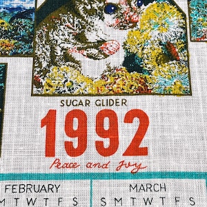 May include: A white fabric calendar from 1992 featuring a printed illustration of a sugar glider surrounded by yellow flowers. The text reads Sugar Glider, 1992, and Peace and Joy in red lettering, with February and March calendar grids visible below.