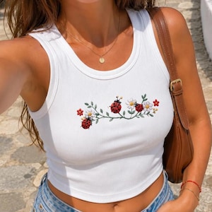 May include: A white ribbed vest top featuring an embroidered design of red ladybugs and small white flowers on a green vine across the chest. The top has a scoop neckline and is paired with blue denim shorts and a brown leather shoulder bag.
