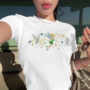 Puede incluir: Una camiseta blanca de manga corta con un estampado gráfico central de un hada con ropa verde y alas translúcidas, rodeada de estrellas amarillas, líneas onduladas, pequeños detalles verdes y una pequeña ilustración de mariposa.