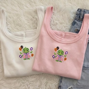 Può includere: Una canotta a coste bianca e una canotta a coste rosa chiaro, entrambe caratterizzate da un ricamo raffigurante un camaleonte verde che indossa un vestito viola e tiene in mano diversi palloncini colorati con facce di zucca e fantasmi.