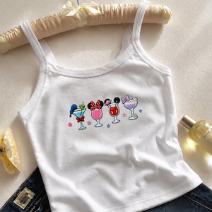 May include: A white sleeveless tank top featuring an embroidered graphic of four colorful cocktail glasses, each stylized with iconic character-inspired details including bows, ears, and hats, arranged horizontally across the center of the chest.