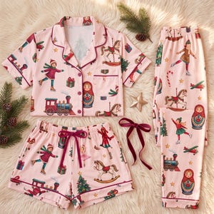 May include: A light pink pajama set featuring a short-sleeve button-down shirt, shorts, and long pants. The fabric has a repeating print of children ice skating, rocking horses, trains, gift boxes, and matryoshka dolls with dark red trim.