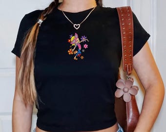 May include: Black crop top with a colorful cartoon graphic of a character surrounded by flowers. The top is paired with blue jeans. A brown purse with a flower charm hangs from the shoulder.