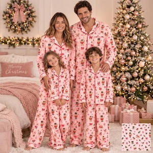 May include: Matching light pink long-sleeve button-down pajama sets featuring a repeating pattern of nutcrackers, rocking horses, and red bows. The pajamas have a collared neckline, front buttons, and long pants with a consistent festive print.