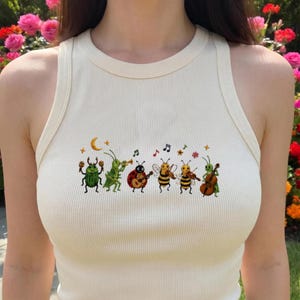 May include: A cream-coloured ribbed tank top featuring a central graphic print of six insects playing musical instruments. The illustration includes a beetle with maracas, a grasshopper with a trumpet, a ladybug with a guitar, and two bees playing violins.
